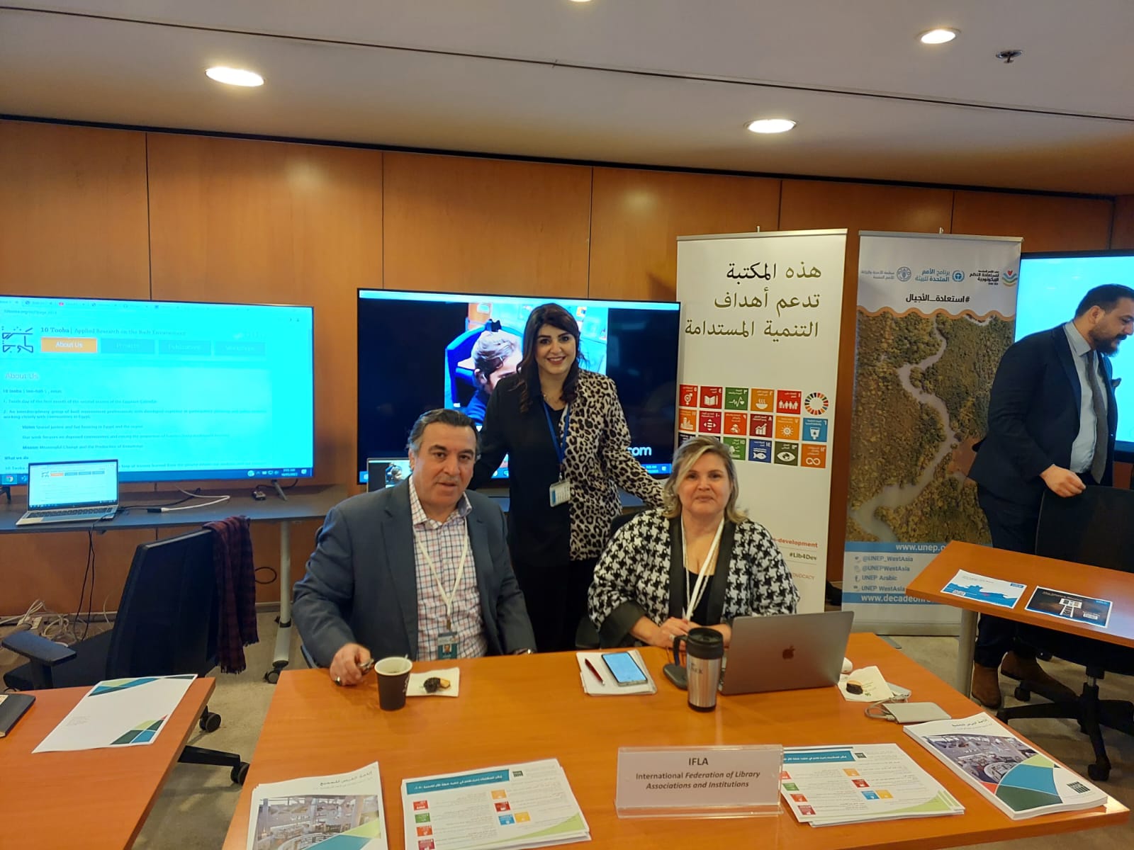 Photo of three people sitting at a table in a conference centre, with banners and screens behind them, and materials on the table in front of them